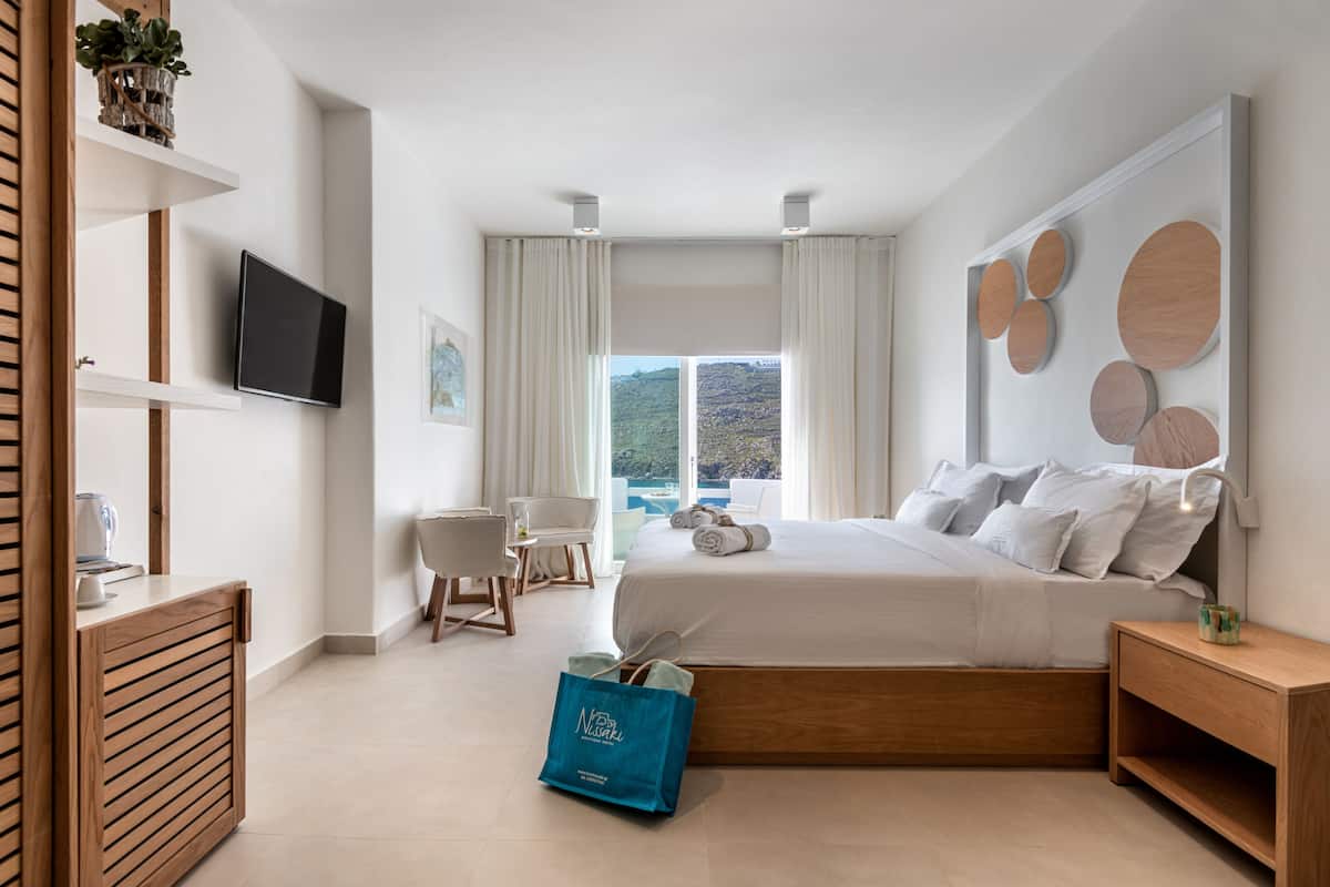 Ein modernes Hotelzimmer mit einem großen Bett, einem an der Wand montierten Fernseher, einem Schreibtisch mit Stuhl und Blick ins Freie durch das Fenster.