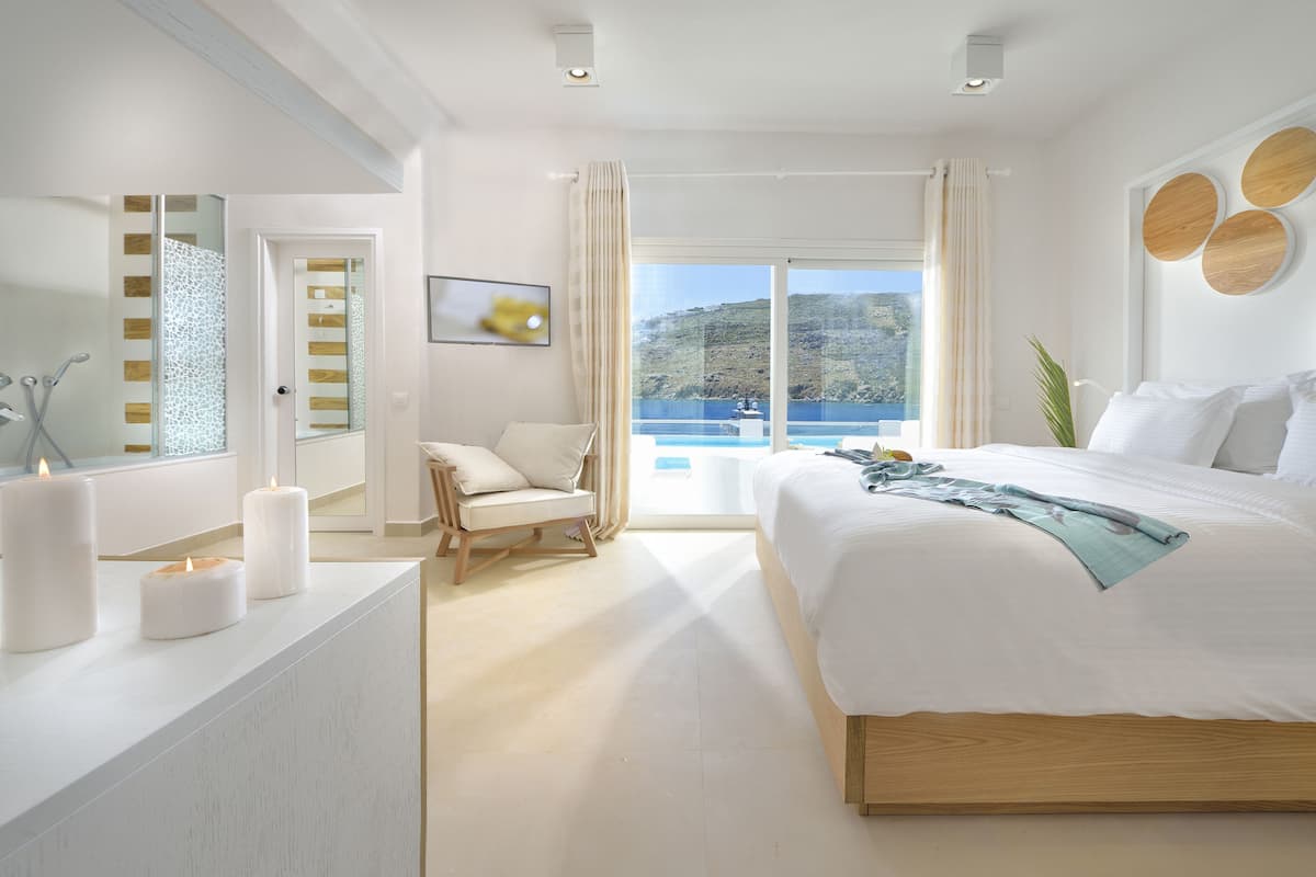 Ein modernes Schlafzimmer mit einem großen Bett, einer Sitzecke mit einem Sessel, einem Schreibtisch mit Kerzen und Meerblick durch große Fenster.