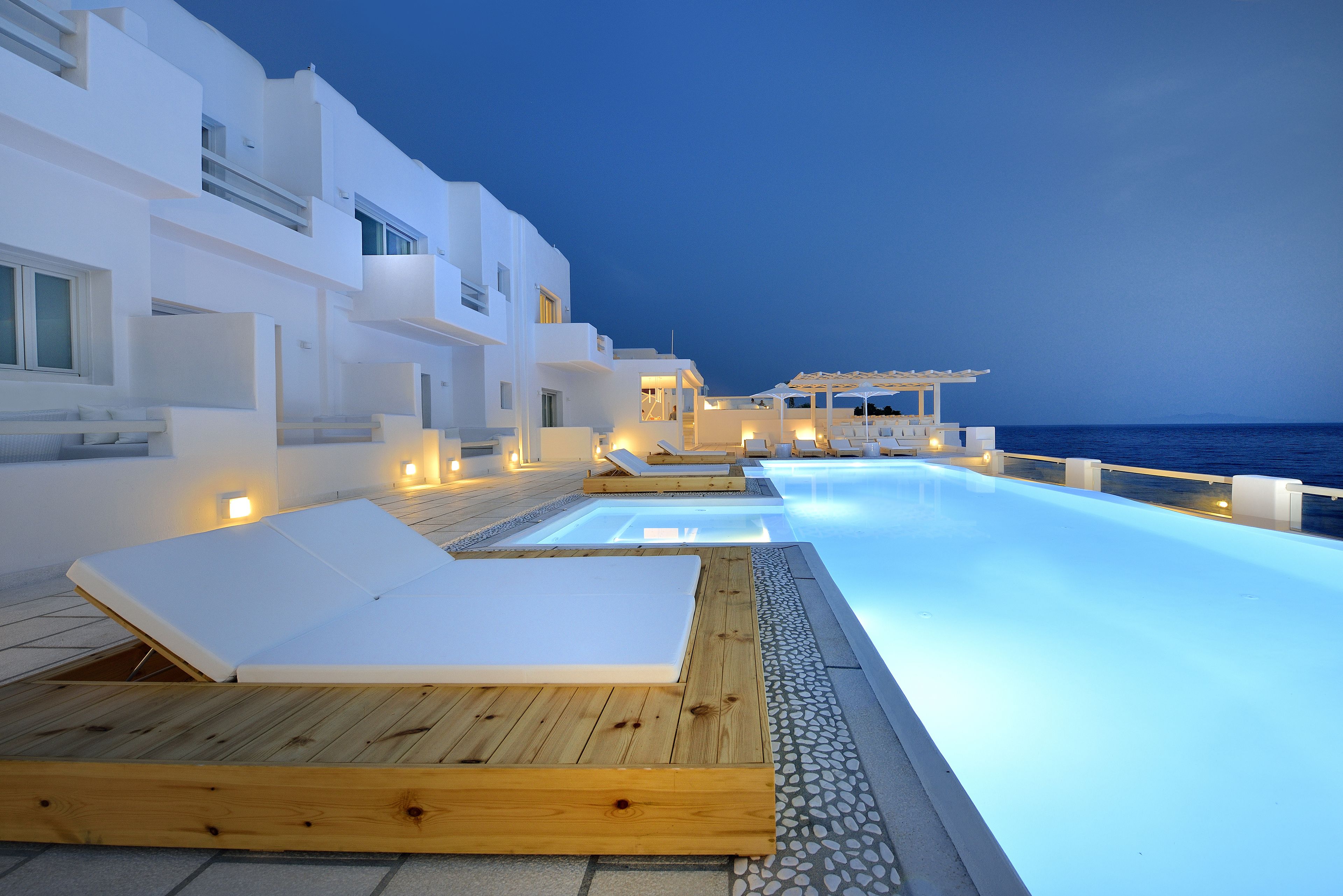 A modern hotel with a infinity pool, wooden loungers, and a view of the sea at dusk.