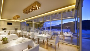A modern dining area with tables set for service, large windows offering a view of the outdoors, and a contemporary ceiling design. - Nissaki Boutique Hotel (Mykonos)