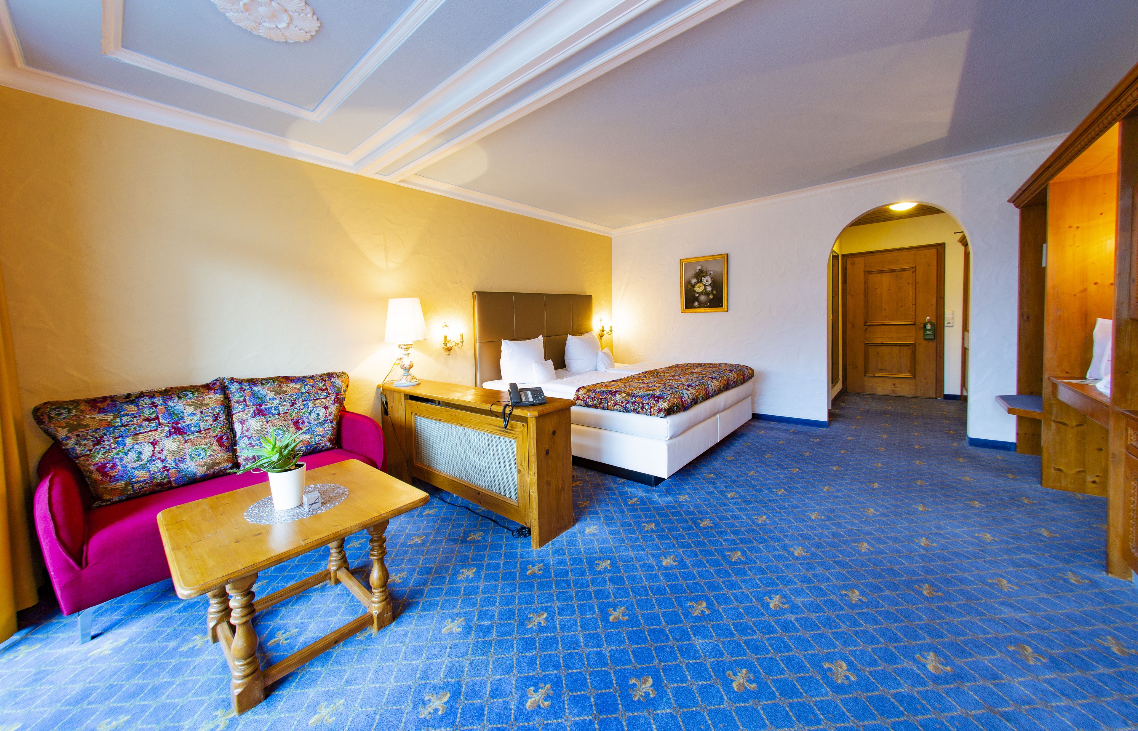 A hotel room with a blue patterned carpet, a pink sofa with colorful cushions, a wooden desk, a white bed with a colorful bedspread, and a wooden door.