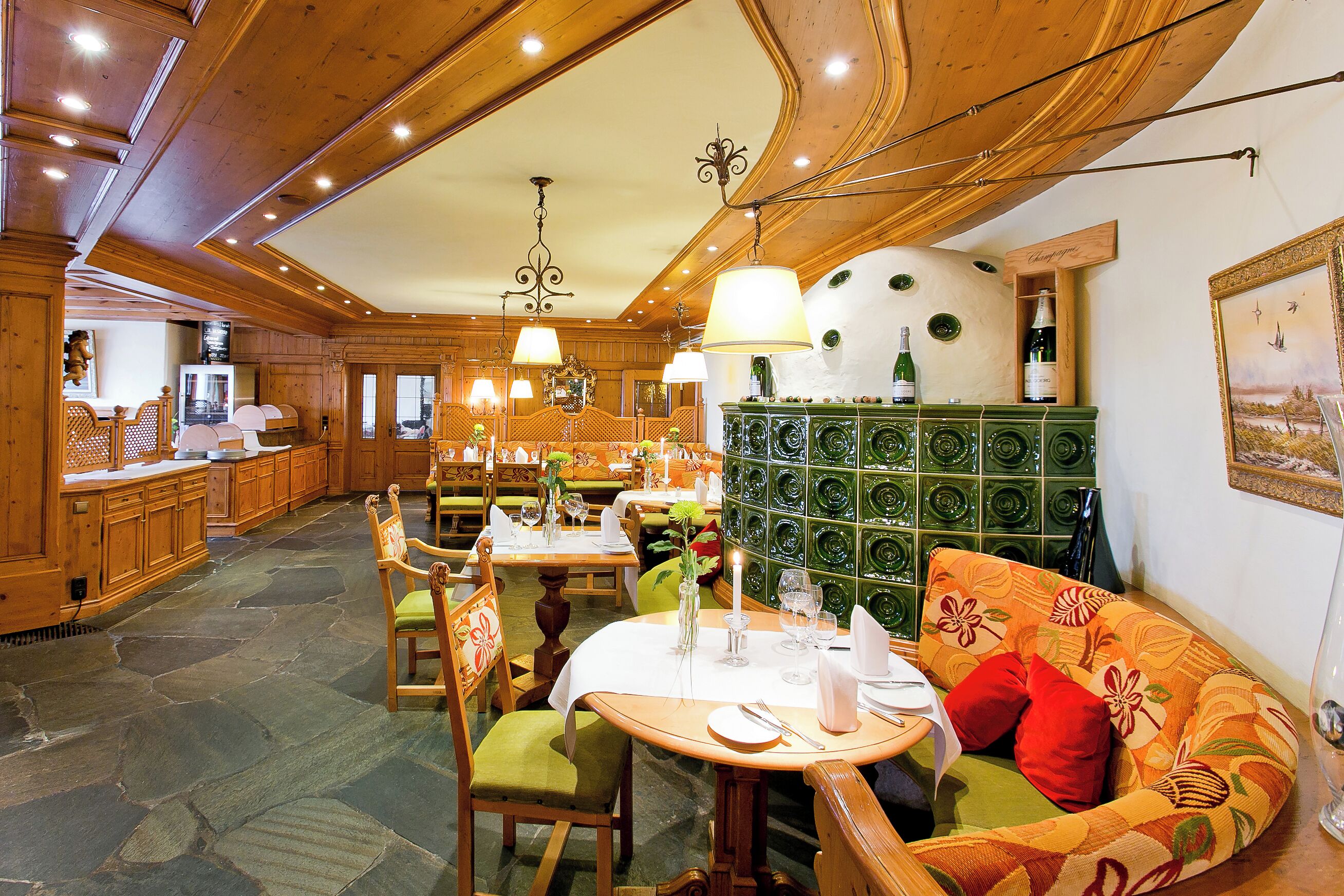 Un restaurante con interiores de madera, suelo de piedra y una pared de mosaico verde. Las mesas están preparadas con manteles blancos y velas.