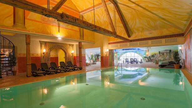 An indoor swimming pool with a wooden ceiling, lounge chairs, and a view of an outdoor area with plants and a fountain. - Hotel Reiterhof Bellevue Spa & Resort (Wirsberg)