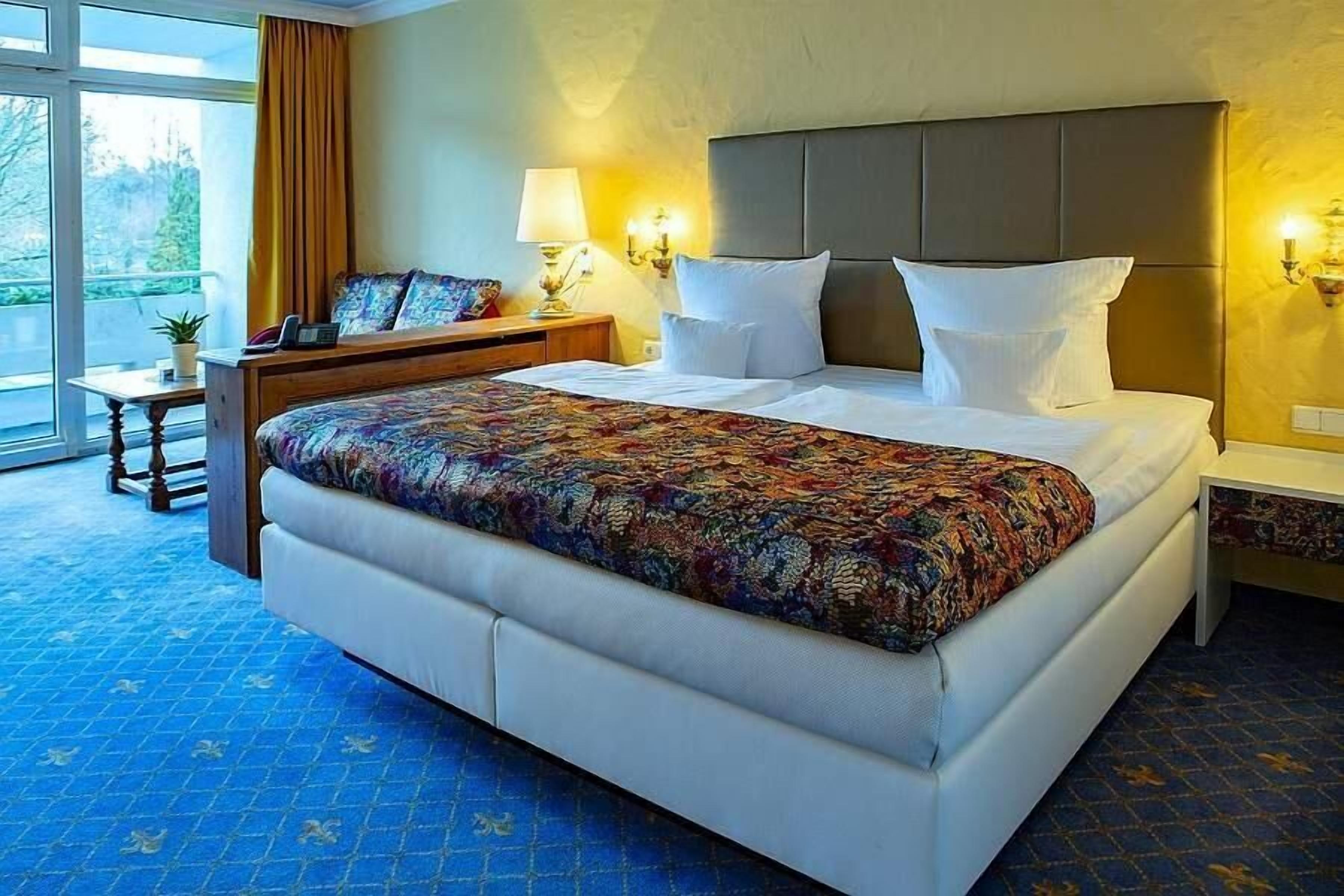 Habitación de hotel con una cama grande, una mesita de noche, una lámpara y vistas al exterior.