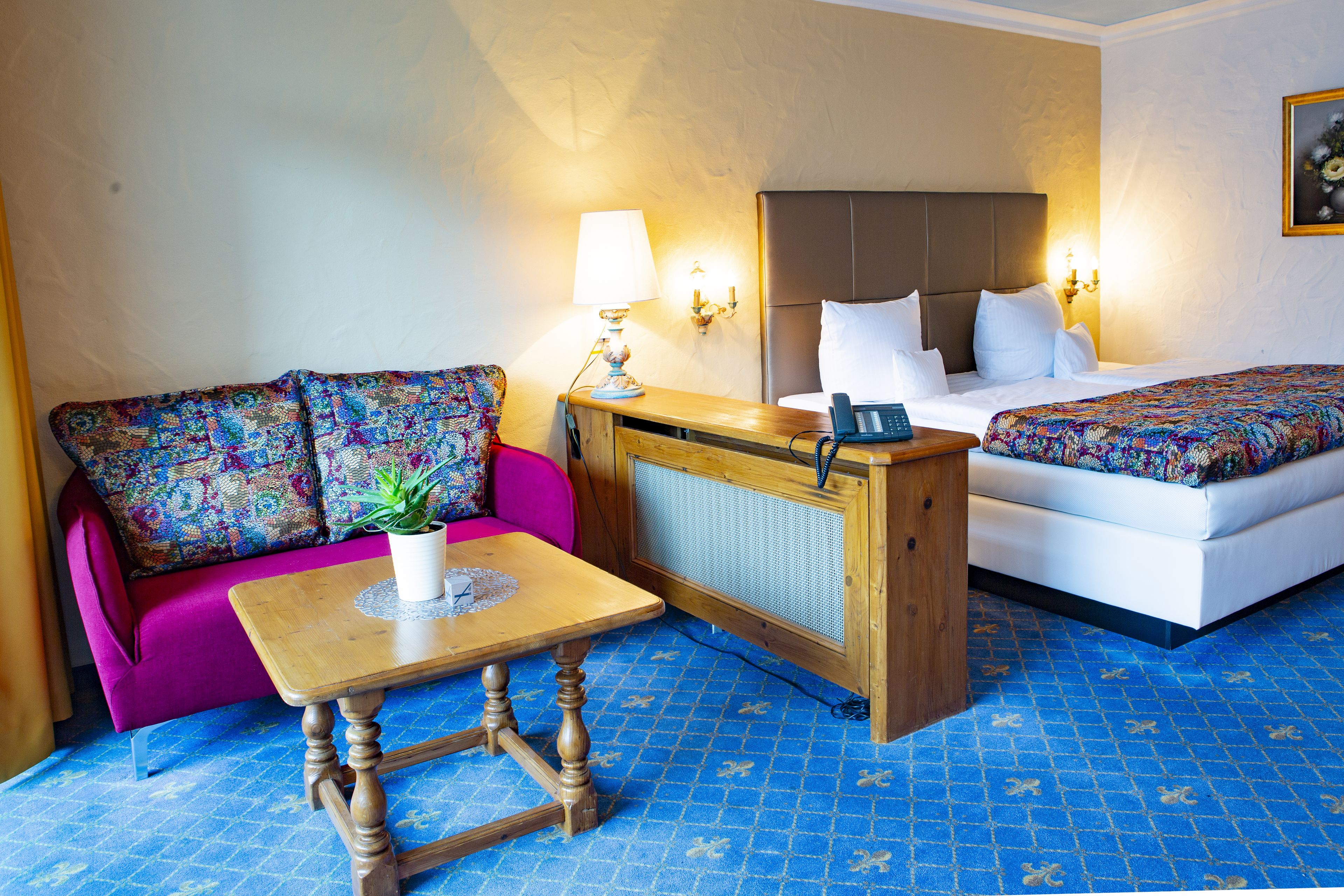 Habitación de hotel con sofá, una pequeña mesa con una planta, cama con colcha colorida y teléfono fijo en la pared.