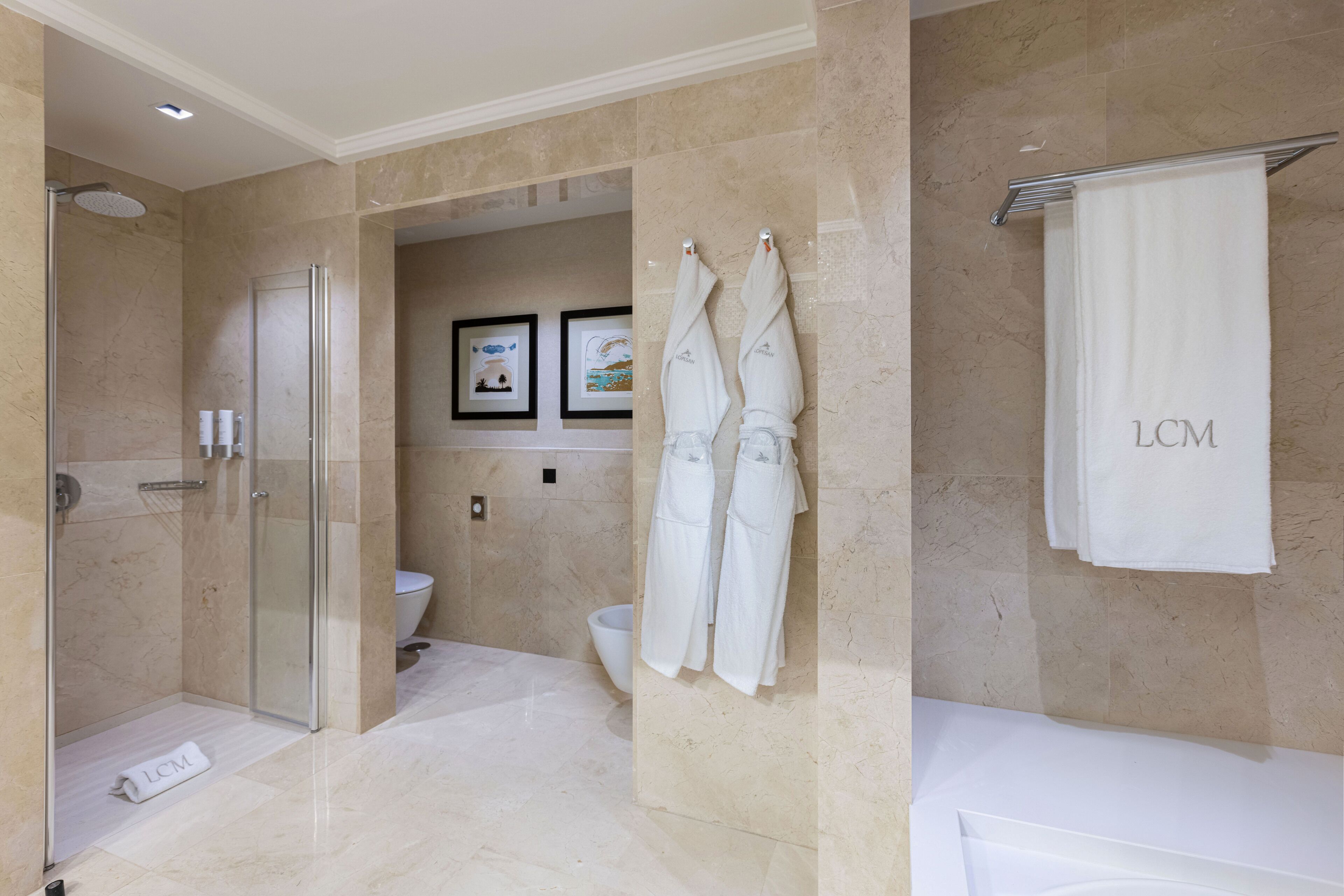 A modern bathroom with a glass-enclosed shower, a toilet, and a bathtub. There are two white towels with 'LCM' on them hanging on the wall.