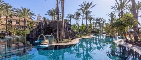 Una amplia piscina exterior con agua cristalina azul, rodeada de palmeras y una cascada artificial de rocas.