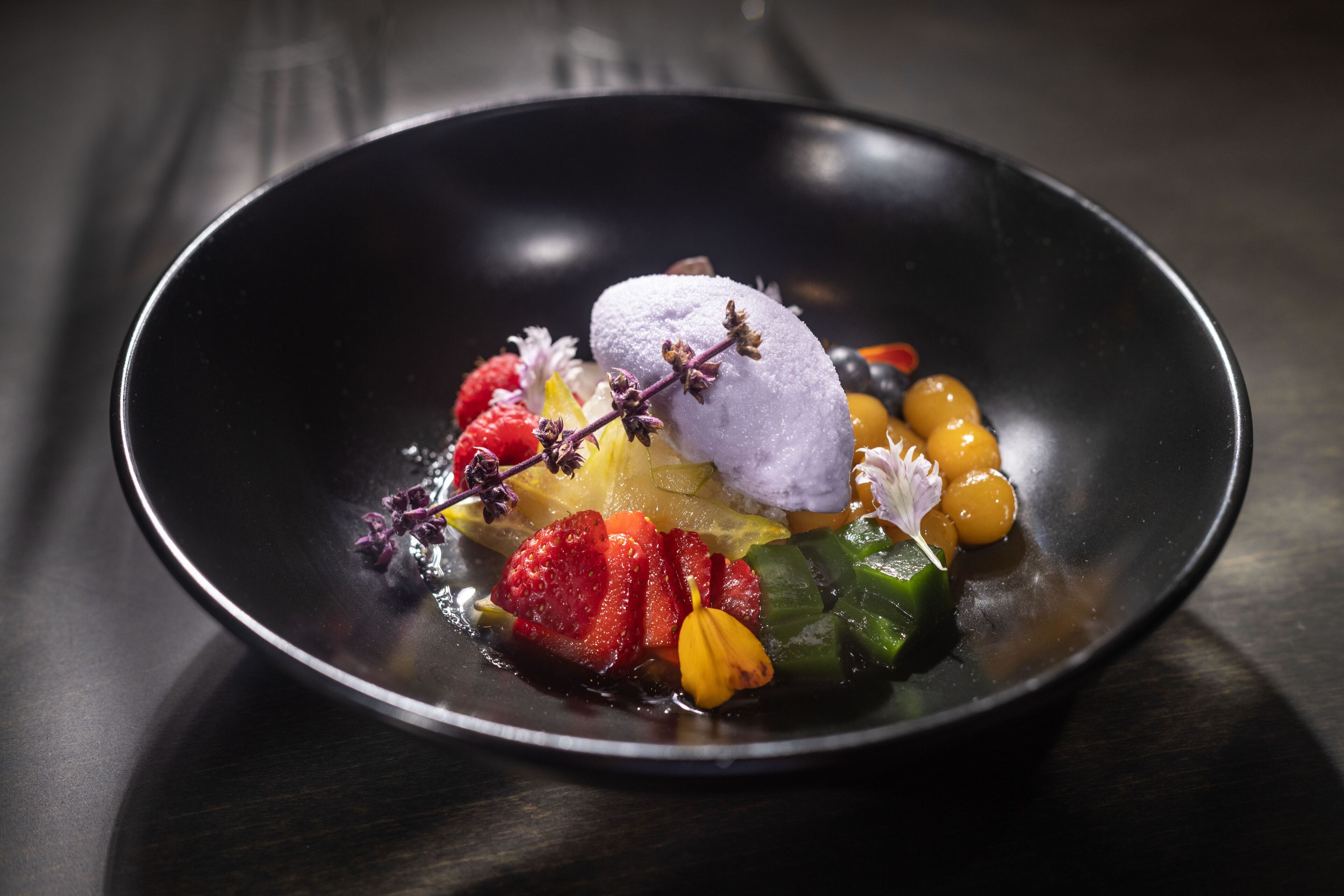Un postre con diversas frutas y una bola de helado sobre un plato negro.