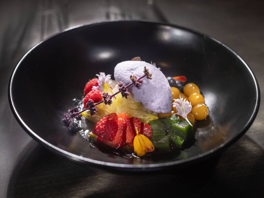 Un postre con diversas frutas y una bola de helado sobre un plato negro.