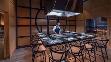 Un restaurante moderno con una mesa central rodeada de taburetes de bar, un chef preparando comida y una decoración interior contemporánea con patrones geométricos en las paredes.