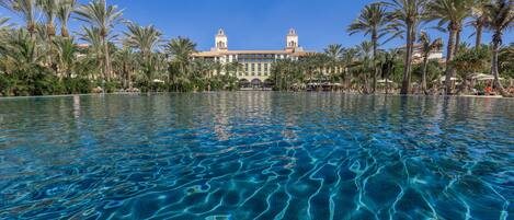 Piscina cristalina con reflejos, rodeada de palmeras y un edificio con dos torres.