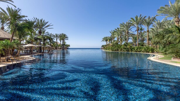 A large infinity pool with clear blue water, surrounded by palm trees and lounge chairs, overlooking a body of water. - Lopesan Costa Meloneras Resort & Spa   (San Bartolomé de Tirajana)