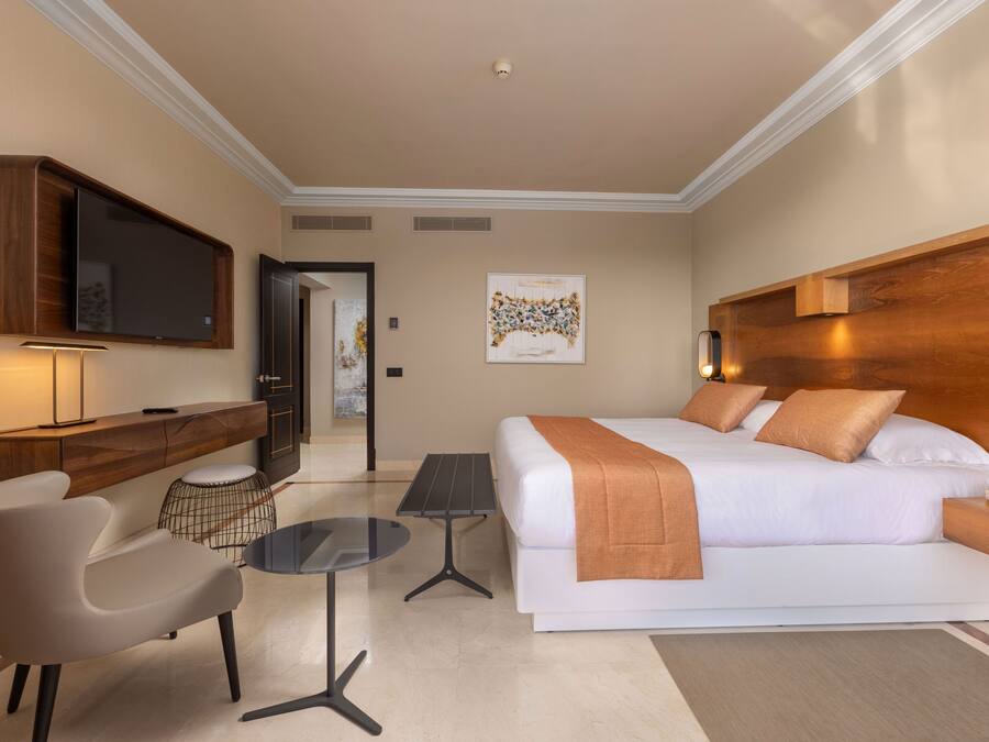 A modern hotel room with a large bed, a desk, a chair, a TV, and a small table.