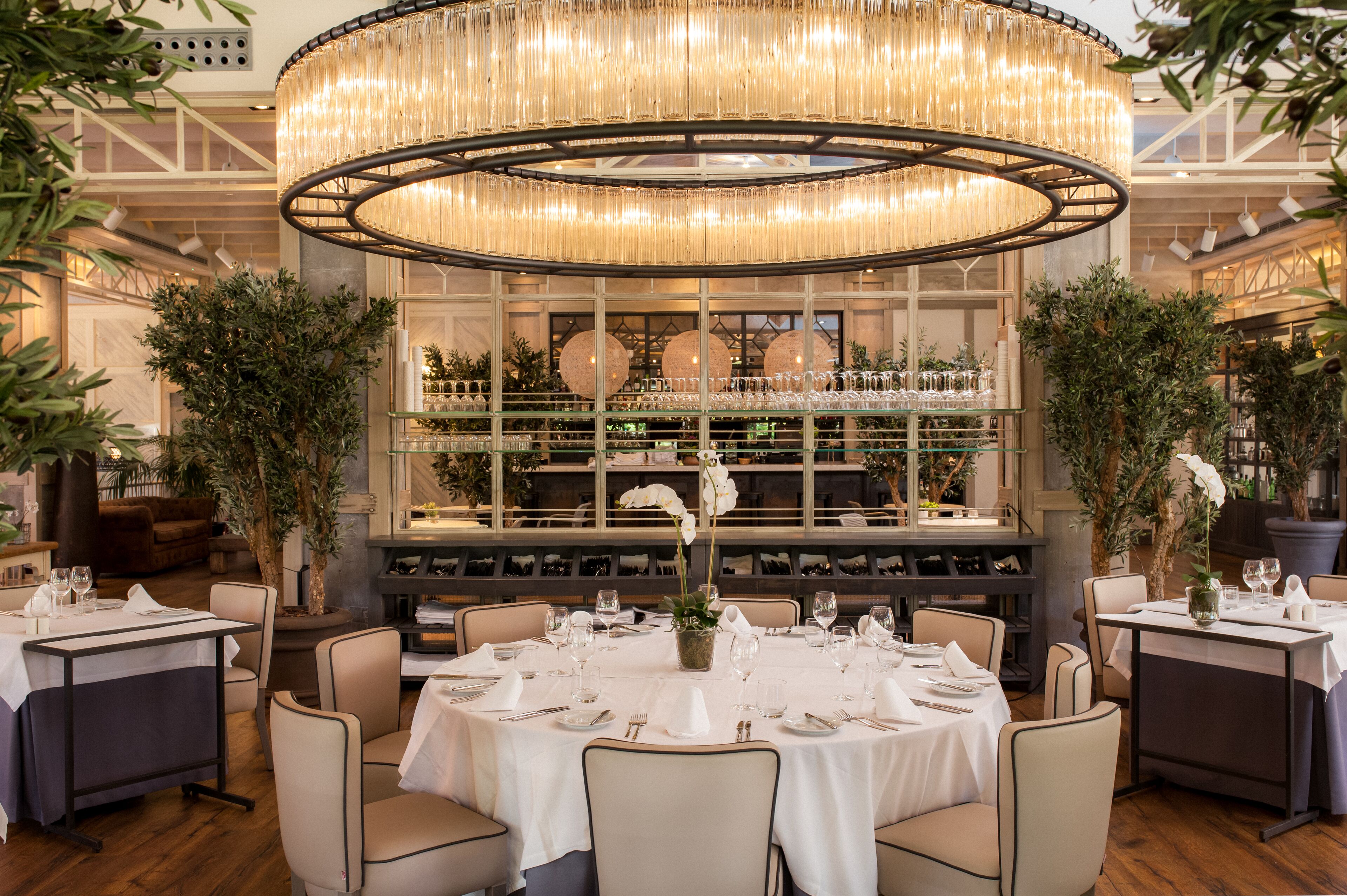 Un restaurante con mesas redondas dispuestas para comer, una araña central y estanterías con cristalería.