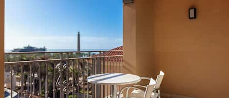 A balcony with a white table and chair, a metal railing, and a clear view of the sea and buildings in the distance.