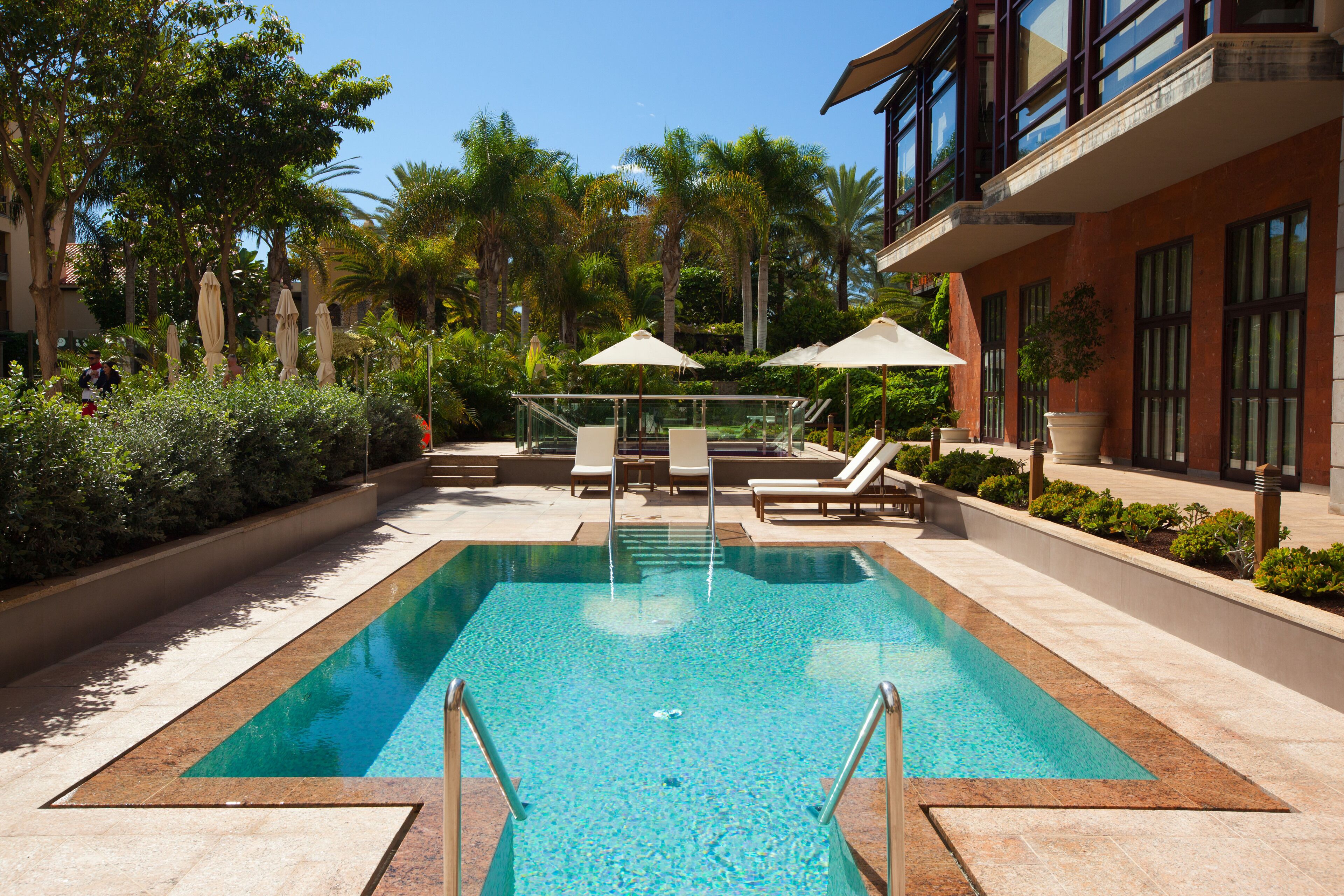A rectangular swimming pool with clear blue water, surrounded by lounge chairs and umbrellas, with a building featuring large windows and balconies in the background.