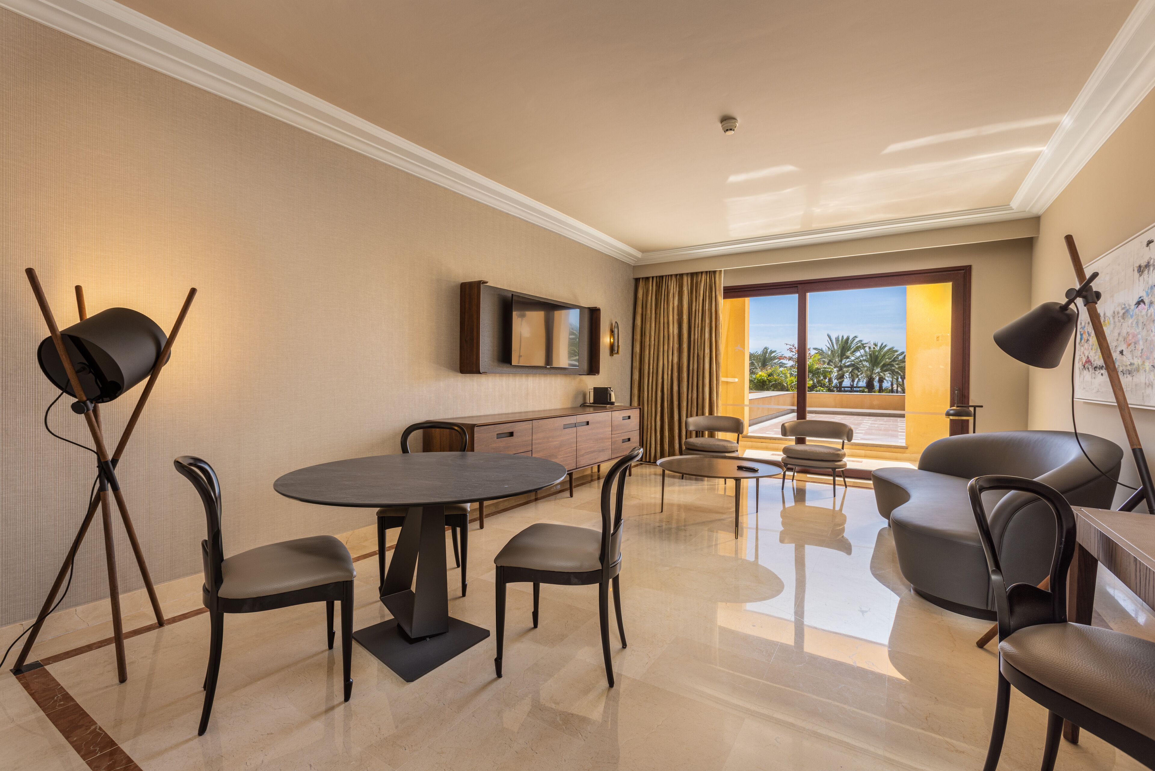 Habitación de hotel moderna con zona de comedor, televisor de pantalla plana en la pared y un amplio ventanal con vistas a palmeras.