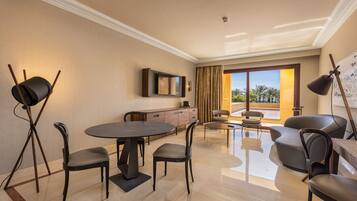 Habitación de hotel moderna con zona de comedor, televisor de pantalla plana en la pared y un amplio ventanal con vistas a palmeras.