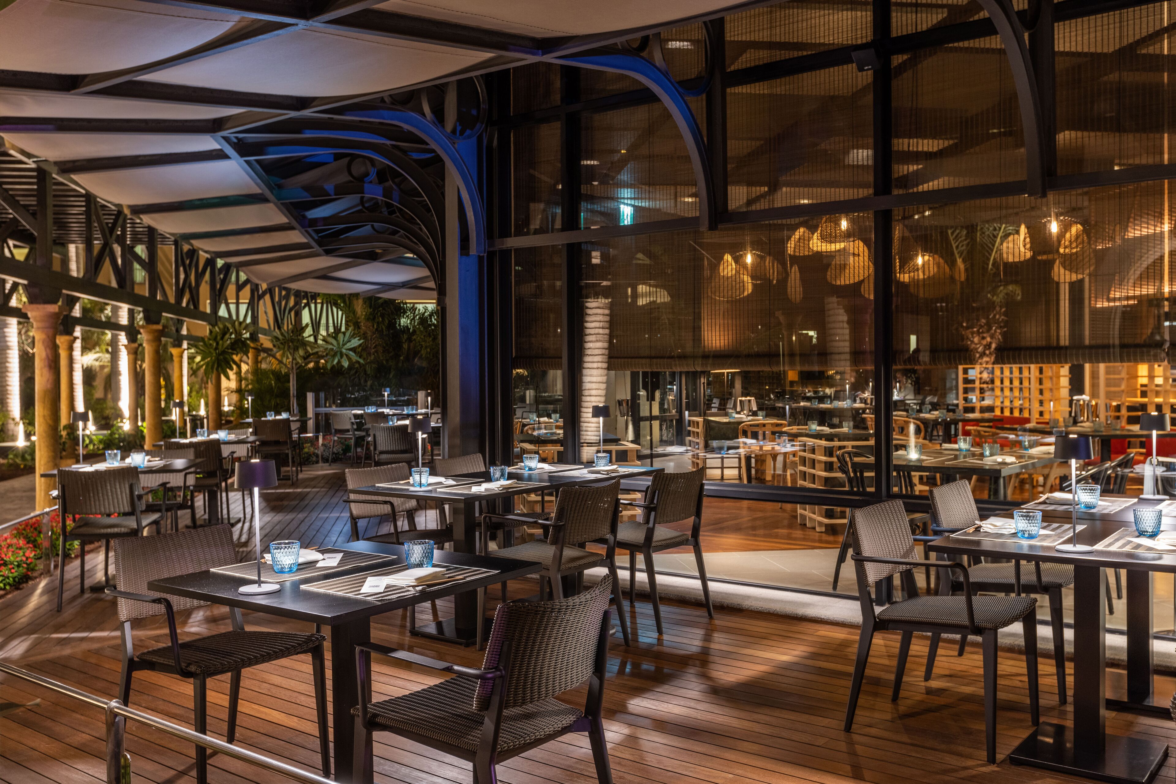 Un área de comedor al aire libre con mesas y sillas, ventanas amplias y un diseño arquitectónico moderno.