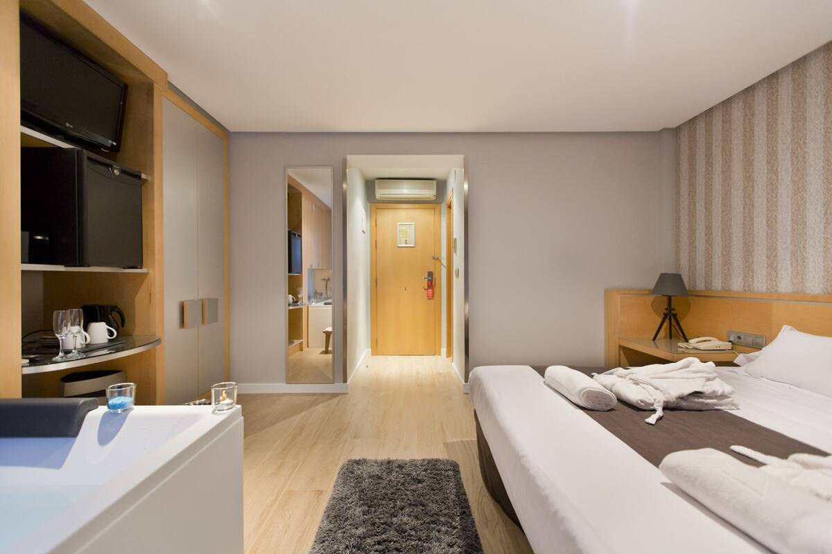 A hotel room with a bed, a desk with a computer, a TV, and a bathroom visible through an open door.