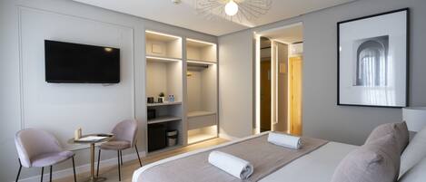 A modern hotel room with a large bed, a television, a desk with two chairs, built-in shelving, and a ceiling light fixture.