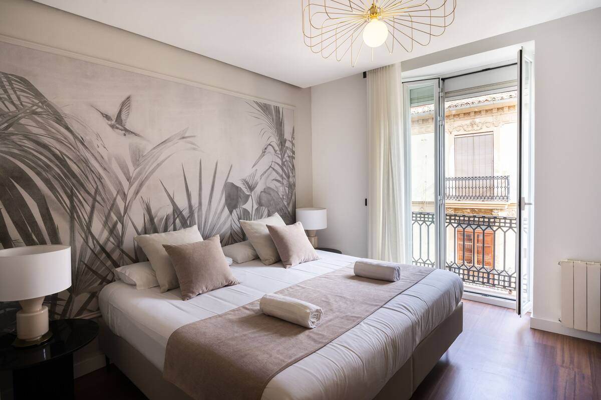 A hotel room with a large bed, two bedside tables, a wall mural, a balcony with a view, and a modern light fixture.