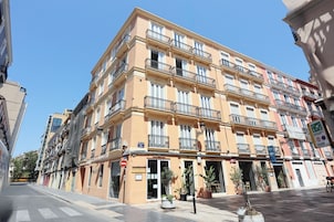 Un hotel de varios pisos con balcones, entrada a nivel de calle y plantas en macetas.