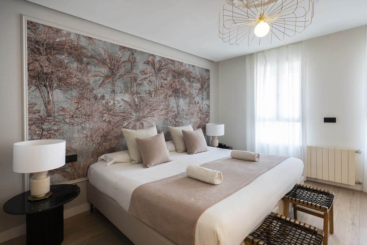 A modern hotel room with a large bed, two bedside tables, a wall mural, a ceiling light fixture, and a window with curtains.
