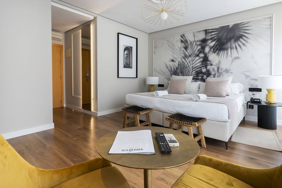 A modern hotel room with a bed, bedside tables, a round table with a remote control, and a framed picture on the wall.