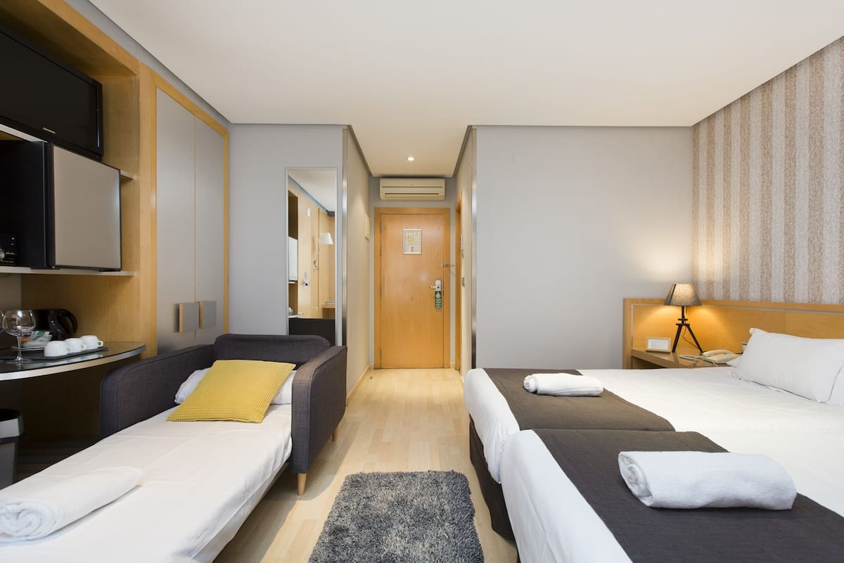 A hotel room with two beds, a desk with a flat-screen TV, a small table with a lamp, and a door leading to another room.