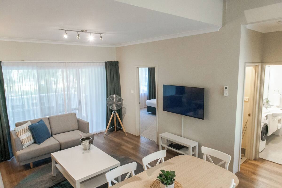 Una sala moderna con sofá, mesa de centro y televisor. Se observa un comedor con mesa y sillas. Un ventilador es visible en una esquina.