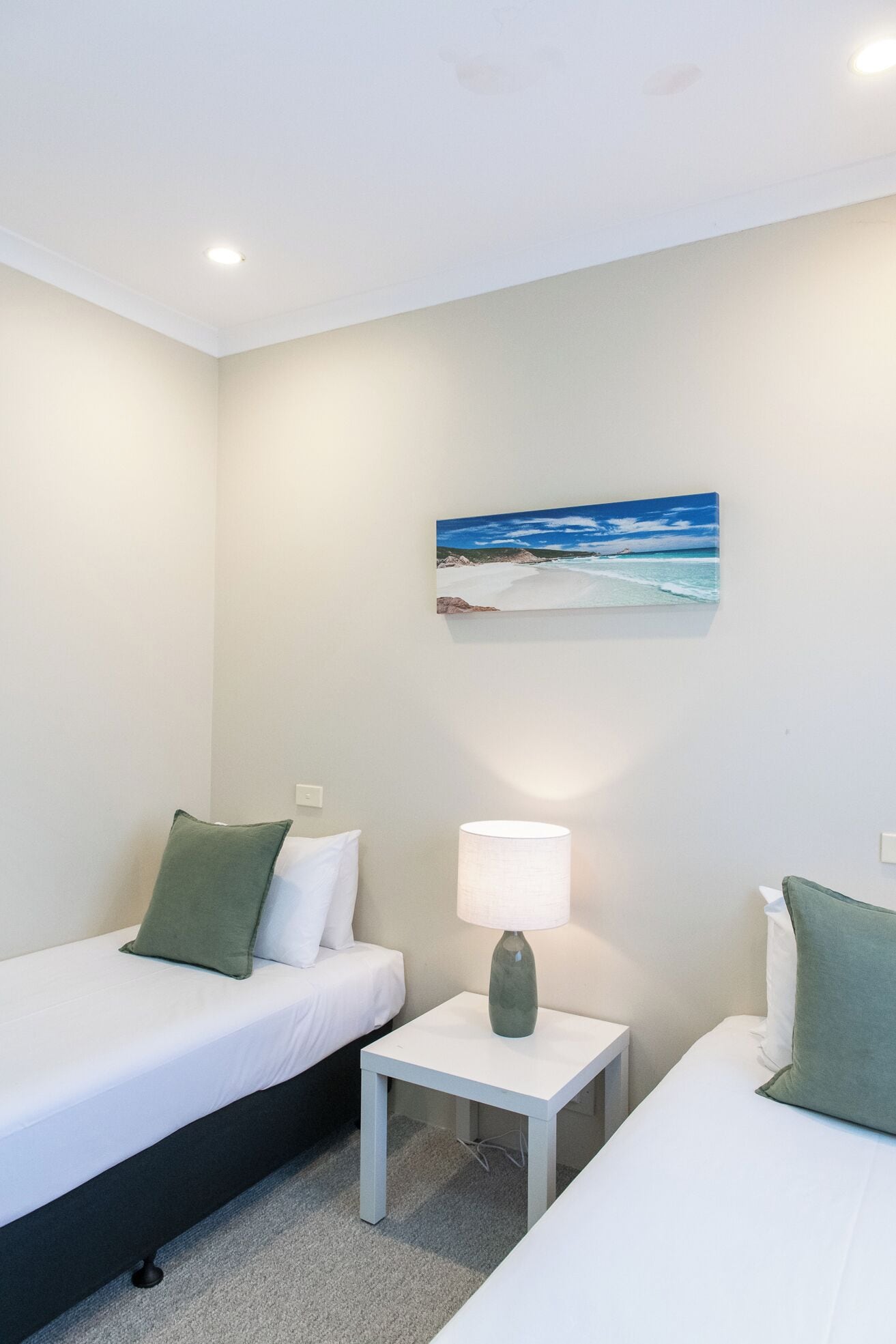 Ein Hotelzimmer mit zwei Einzelbetten, einem kleinen Nachttisch mit Lampe und einem Bild einer Strandansicht an der Wand.