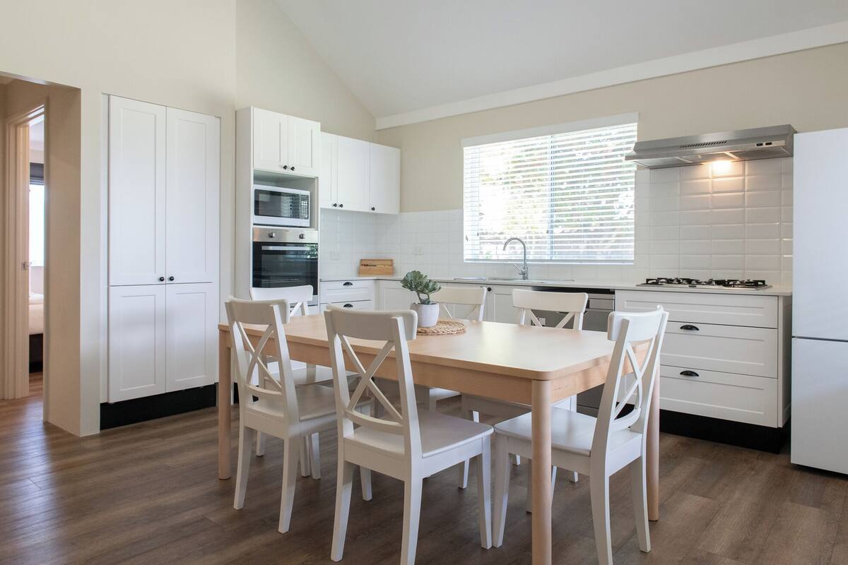 Una cocina moderna con armarios blancos, una mesa de comedor de madera y sillas. Hay una ventana con persianas, un horno empotrado y una estufa.