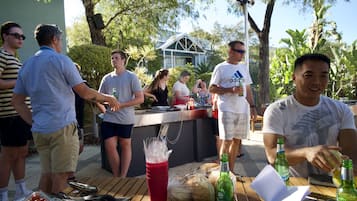 A group of people gathered around a wooden table with food and drinks, under a shaded area with trees.