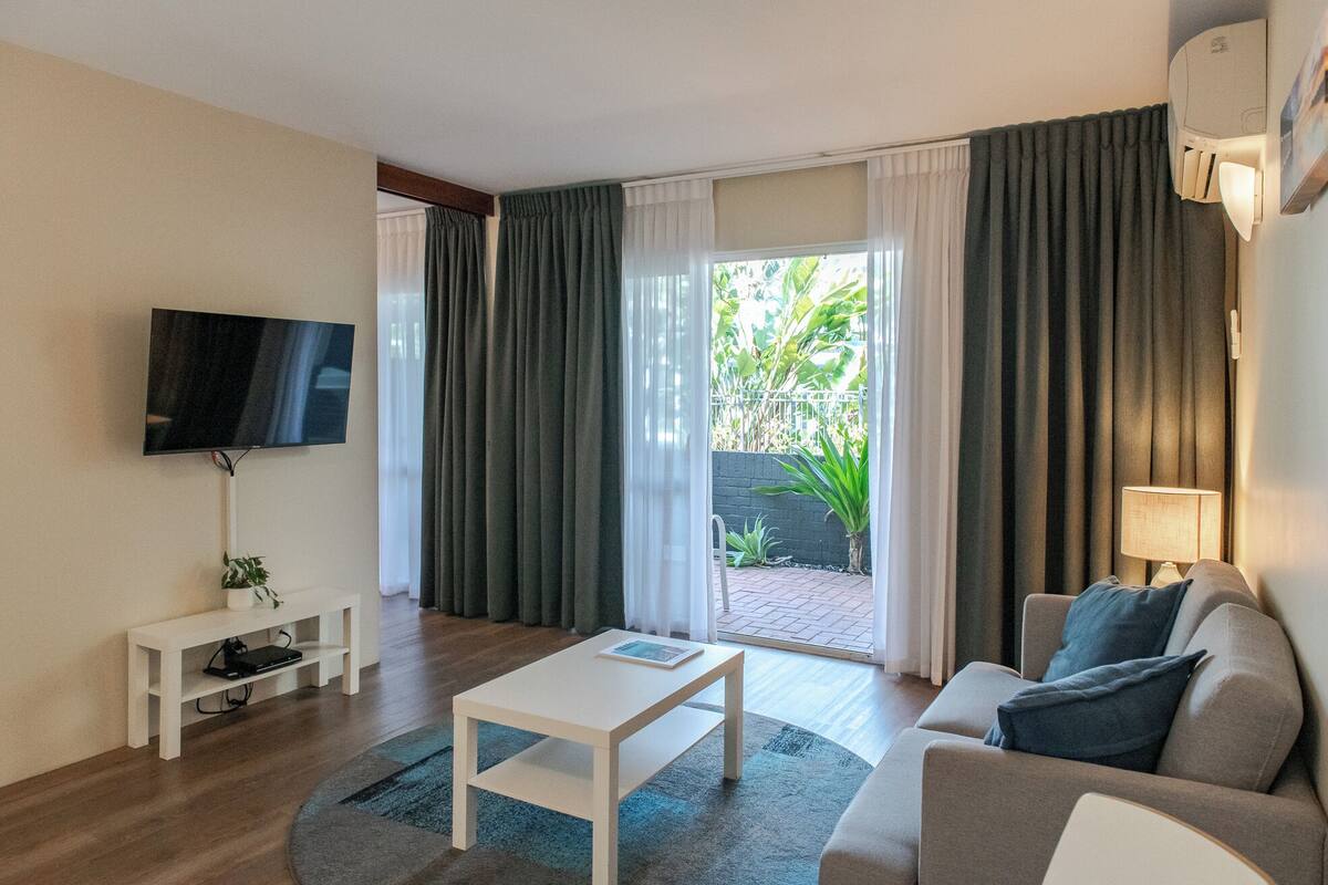 Una sala de estar moderna con un televisor de pantalla plana, una mesa de centro blanca, un sofá gris con cojines azules y una mesita auxiliar blanca.