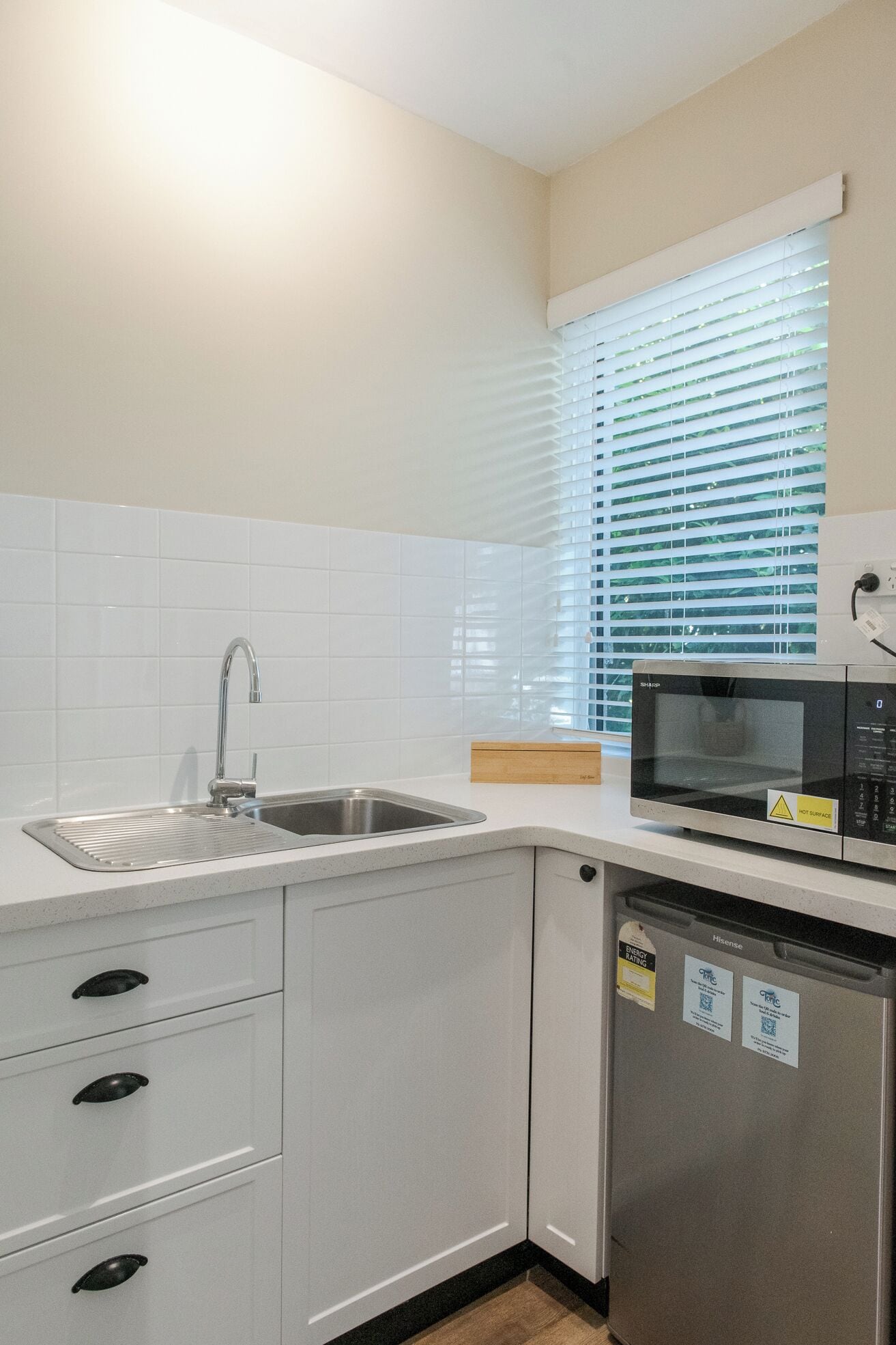 A kitchen with white cabinets, a stainless steel sink, a microwave, and a window with blinds.