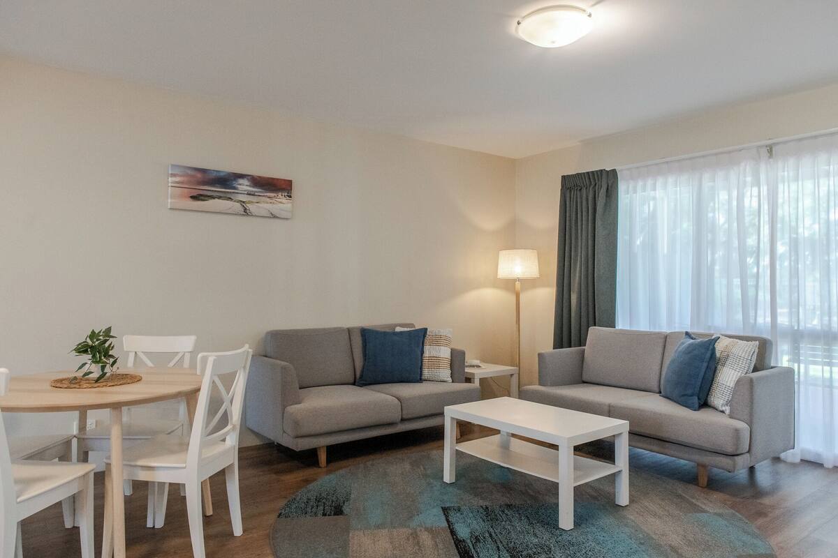 Una sala de estar moderna con un sofá gris, una mesa de centro blanca y un comedor con una mesa pequeña y sillas. Hay un cuadro en la pared y una lámpara sobre una mesita auxiliar.