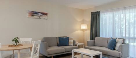 A modern living room with a grey sofa, white coffee table, and a dining area with a small table and chairs. A painting is on the wall, and there is a lamp on a side table.