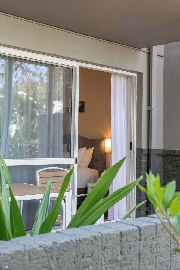 A room with a bed, a bedside table, and a chair. There is a window with curtains, and a plant is in the foreground.