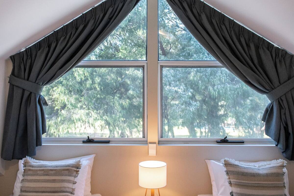 Un dormitorio con dos camas, un ventilador de techo, una ventana con cortinas y una lámpara de noche.