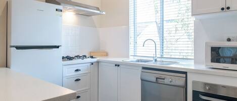 Uma cozinha moderna com armários brancos, lava-louças de aço inoxidável, forno embutido, micro-ondas e uma janela com persianas.