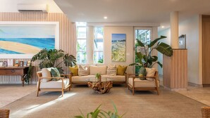Een moderne woonkamer met een bank, fauteuils en een salontafel. Er zijn grote ramen, een schilderij van een strand en een plant.