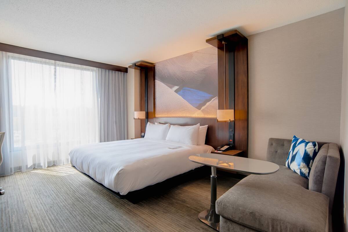 A hotel room with a large bed, a sofa, a small table, and a nightstand with a lamp.