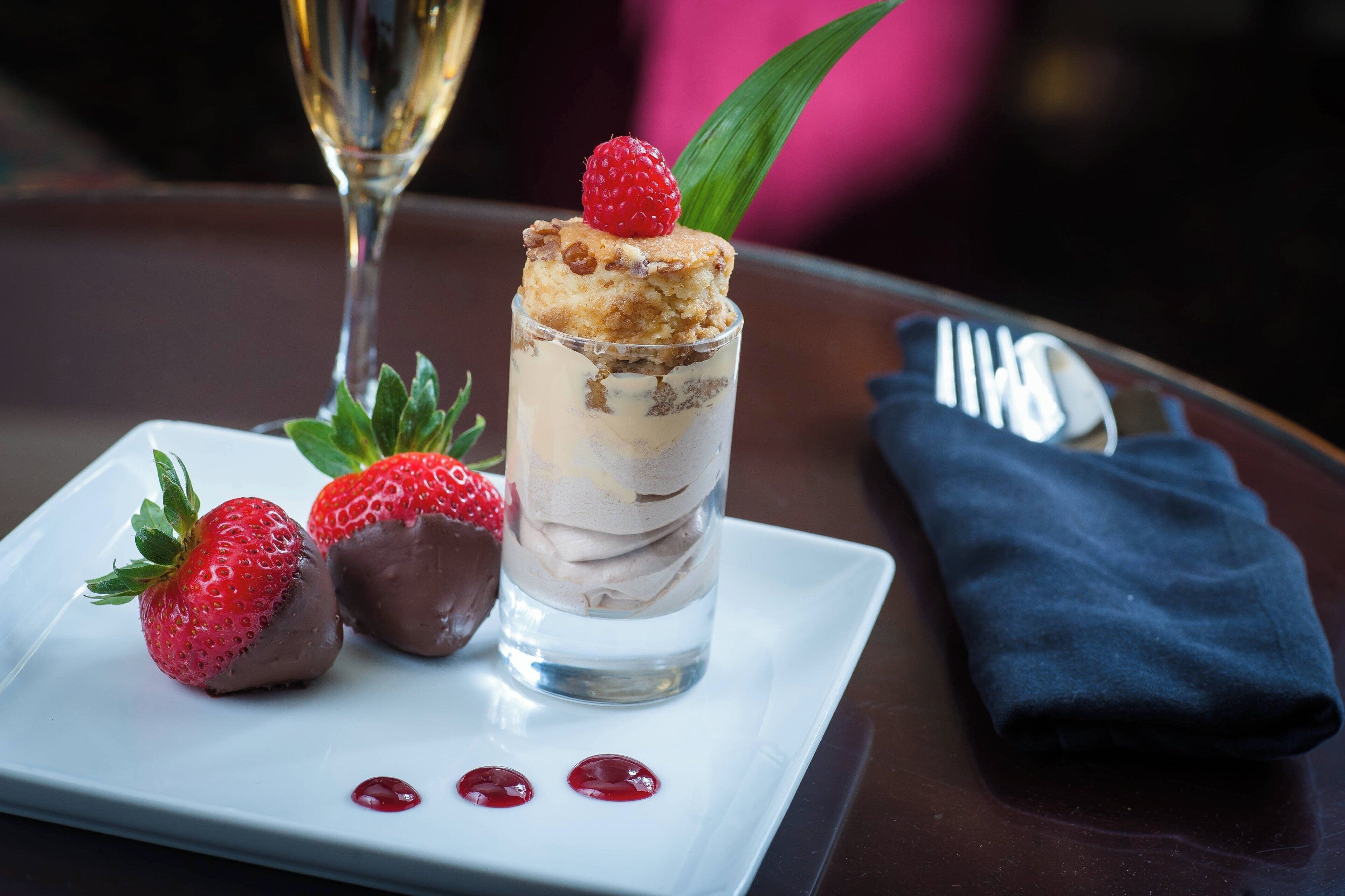Un dessert aux fraises, agrémenté d’une fraise enrobée de chocolat, présenté sur une assiette blanche, accompagné d’un verre contenant un dessert à plusieurs couches et d’une serviette pliée.