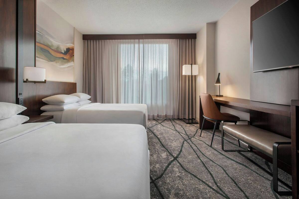A hotel room with two beds, a desk, a chair, a TV, and a large window with curtains.