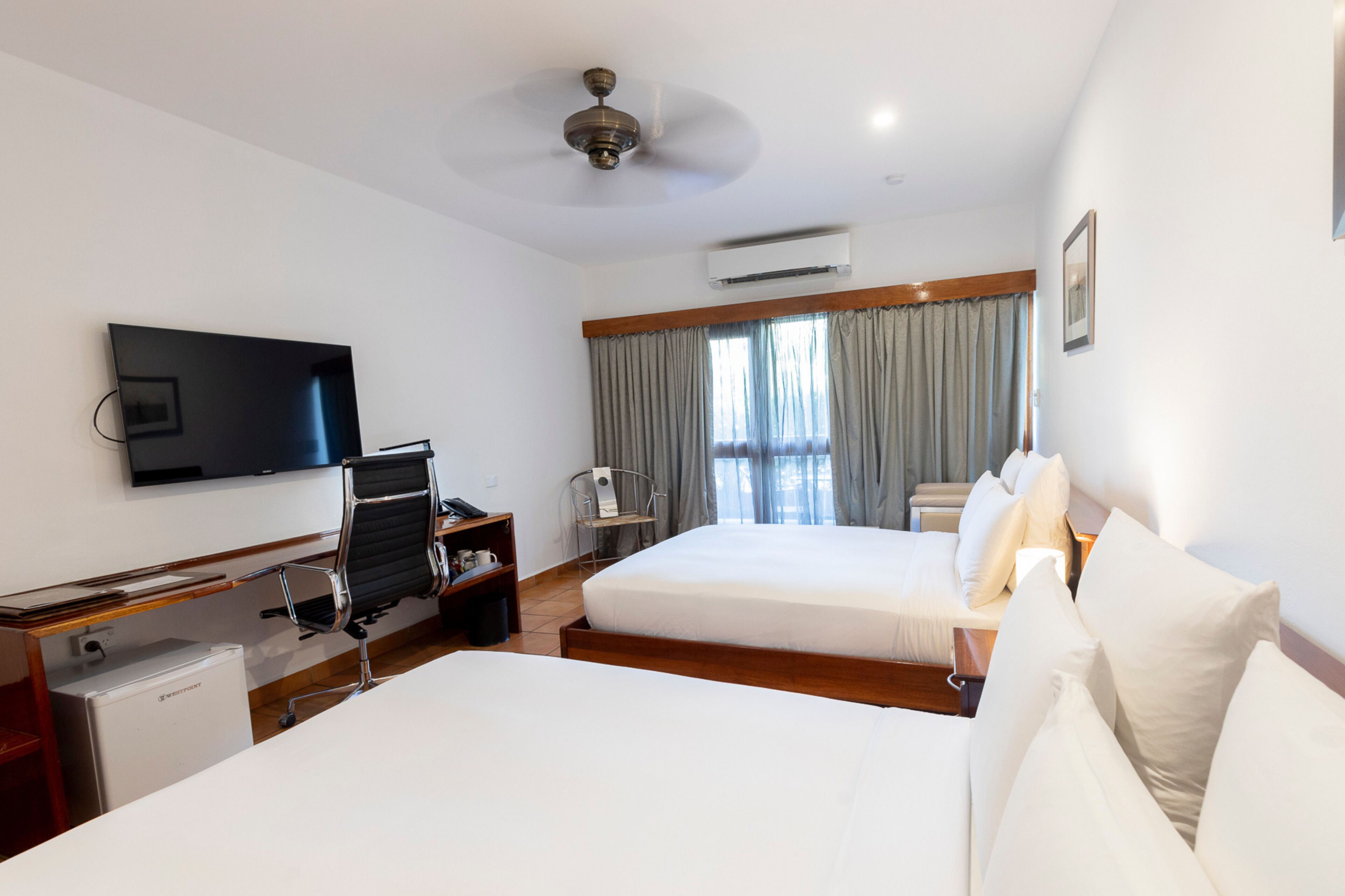 A hotel room with two beds, a desk, a chair, a television, and an air conditioning unit.