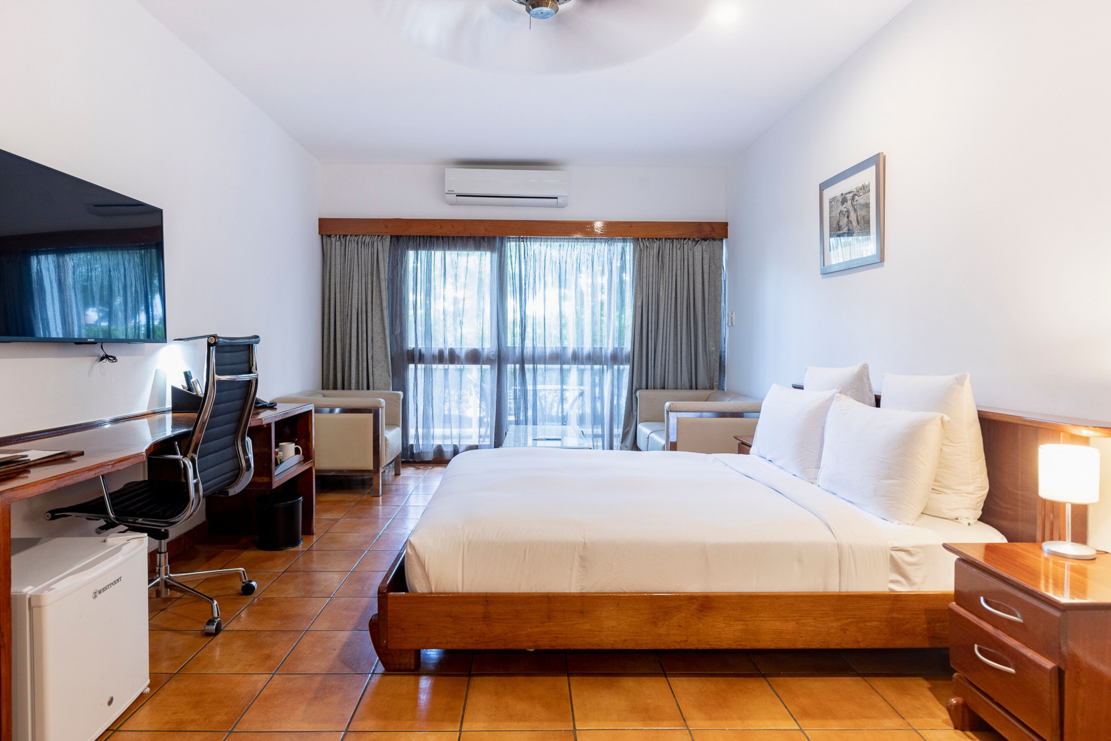 A hotel room with a bed, desk, chair, TV, and air conditioning unit.