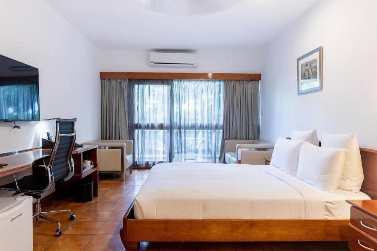 A hotel room with a bed, desk, chair, TV, air conditioning unit, and a window with curtains.