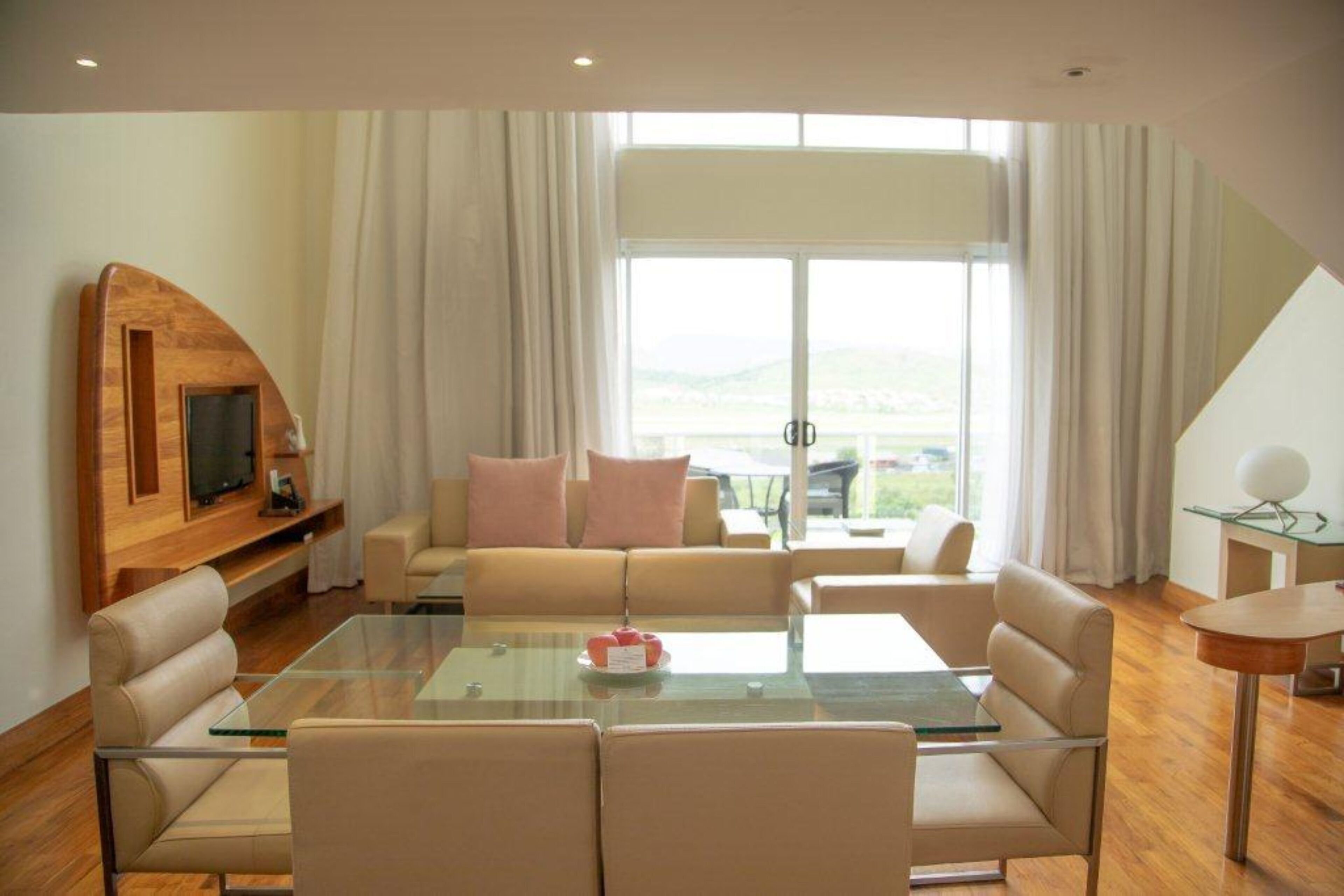 A modern living room with a glass coffee table, beige sofas, a flat-screen TV on a wooden shelf, and a large window with curtains.
