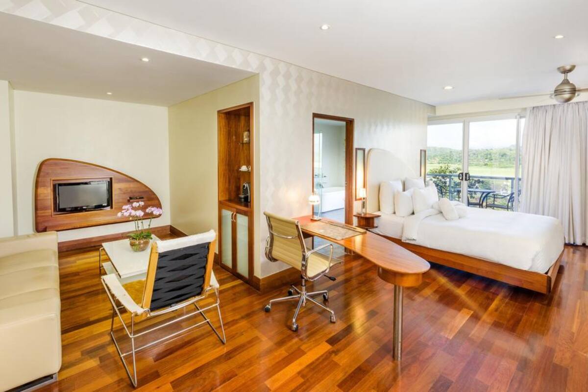 A modern hotel room with a wooden floor, a bed, a desk with a chair, a sofa, a TV, and a balcony with a view.